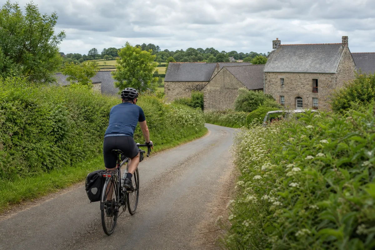 Un cycliste sur une petite route entre haies et bâtiments ruraux en pierre